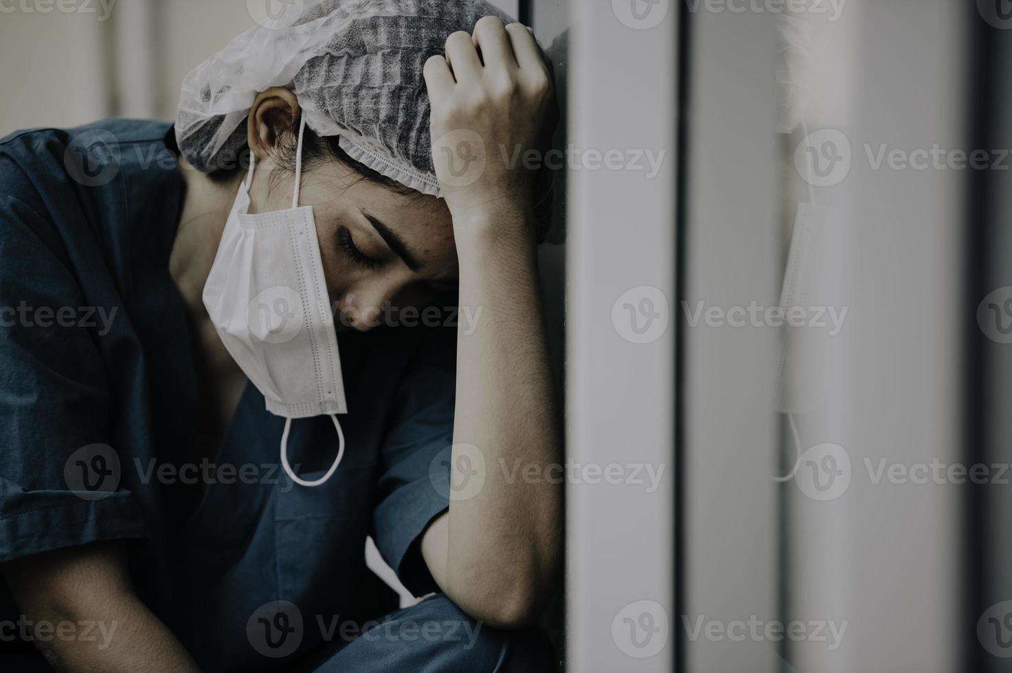 Tired depressed female asian scrub nurse wears face mask blue uniform sits on hospital floor,Young woman doctor stressed from hard work photo