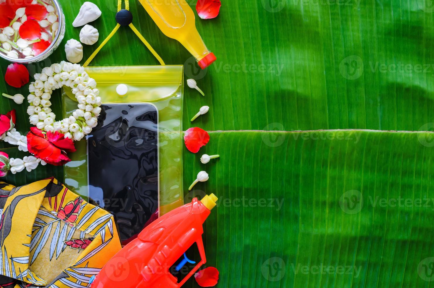 Thailand songkran festival background with smartphone in waterproof case, water gun, shirt, garland and scented water on banana leaf background. photo