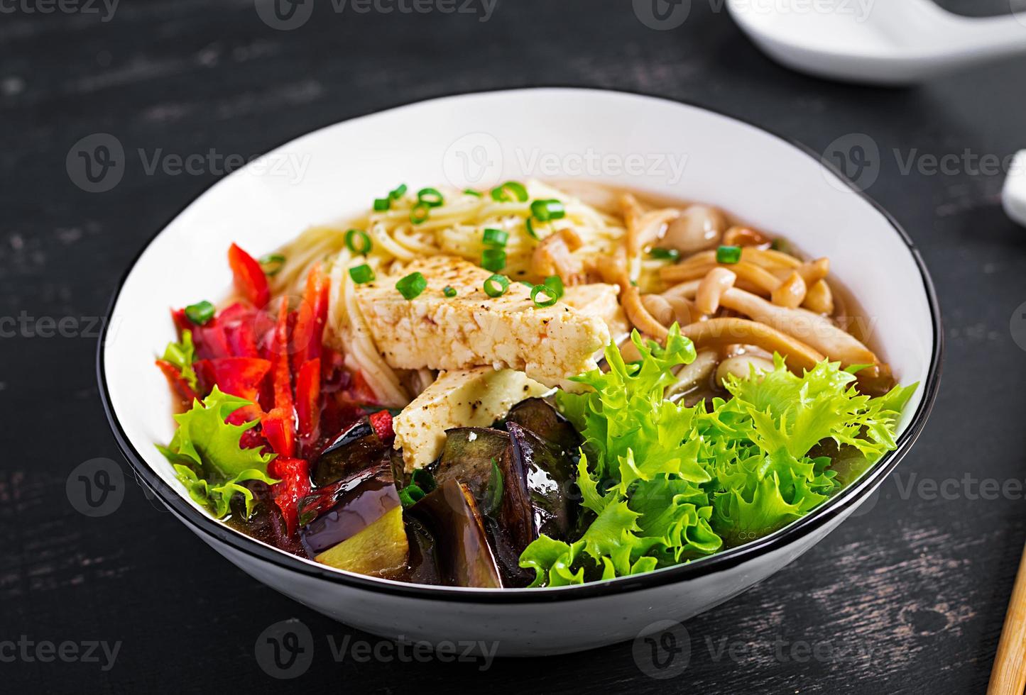 Asian vegan noodles soup with tofu cheese, shimeji mushrooms and fried eggplant in bowl on dark background. photo