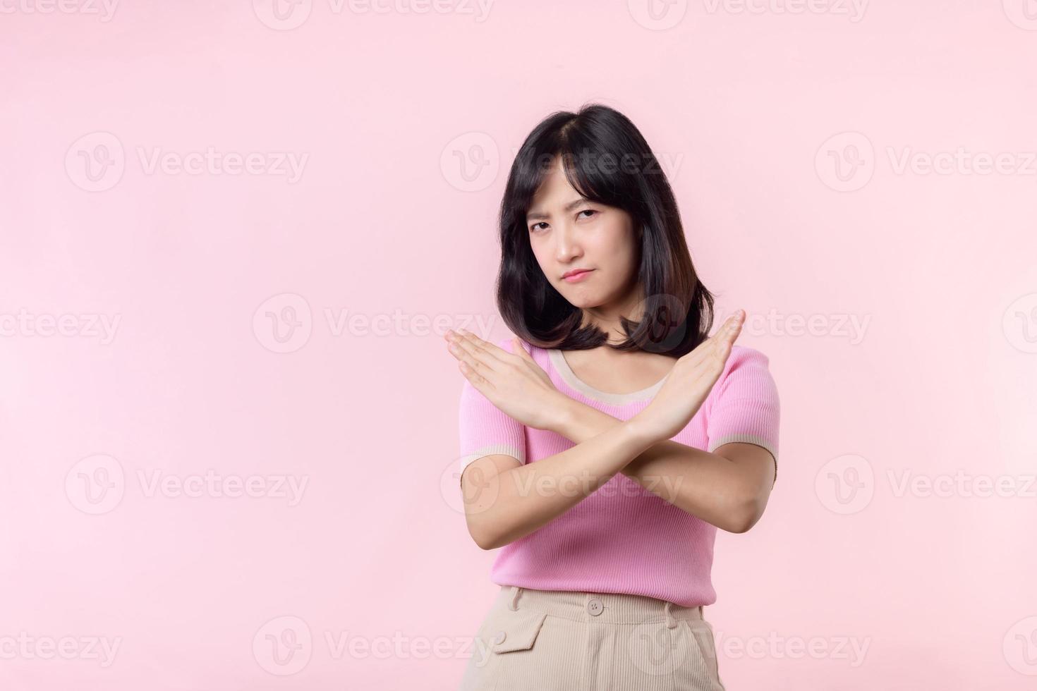 portrait serious young asian woman with cross arm gesture showing stop, no, wrong, denial, rejection sign isolated on pink pastel studio background. deny and negative expression symbol concept. photo