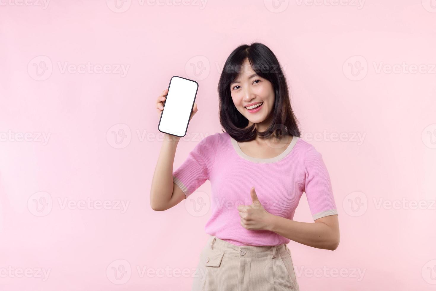 retrato hermosa joven asiático mujer contento sonrisa demostración teléfono inteligente pantalla con señalando dedo mano gesto a móvil teléfono aislado en rosado pastel estudio antecedentes. blanco pantalla teléfono inteligente concepto foto