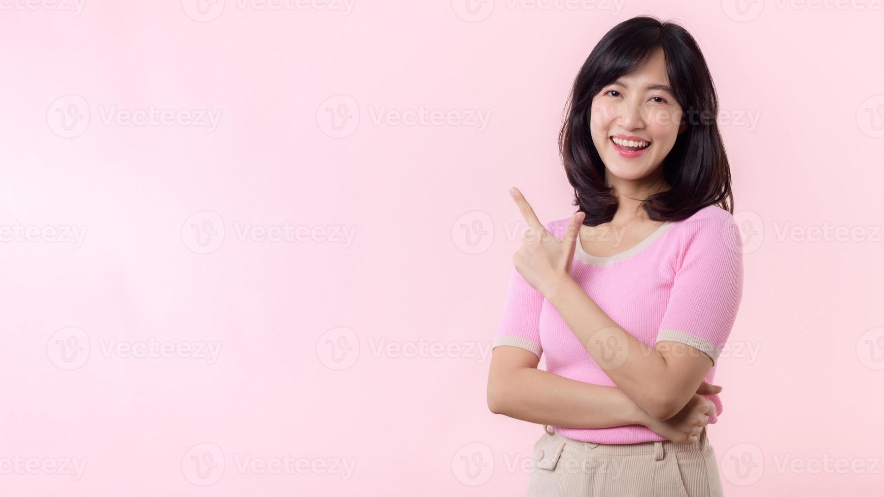 Portrait young beautiful asian woman happy smile pointing finger and hand to showing on copy space isolated on pink background. Attractive female person gesture attention to this promotion. photo