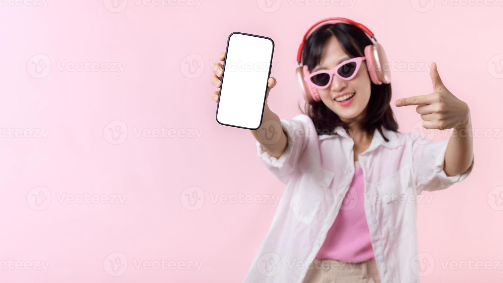 Happy cheerful smiling asian woman with wireless earphones showing blank screen mobile phone or new smartphone music application advertisement mockup isolated on pink studio background. photo