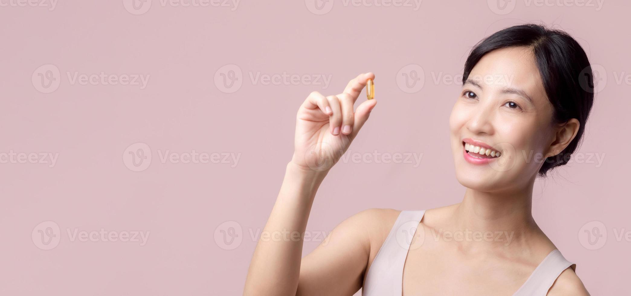 Portrait young asian woman happy smile face with vitamin nourishment pill. Pretty cute girl female person holding health capsule supplement skin care isolated on pink background. Medication concept. photo