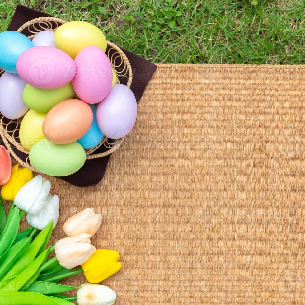 Happy Easter holiday greeting card concept. Colorful Easter Eggs and spring flowers on Papyrus rattan weave texture background. Flat lay, top view, copy space. photo