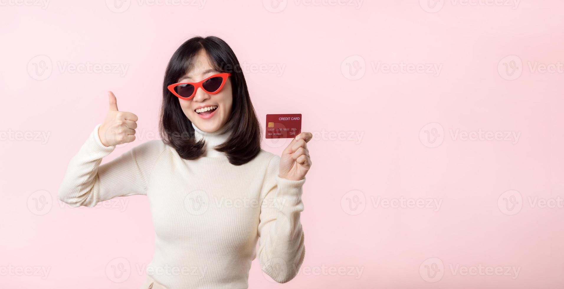 Fascinating fun joyful young woman of Asian ethnicity 20s years old with wear sunglasses wears white shirt hold in hand credit bank card isolated on plain pastel light pink background studio portrait. photo