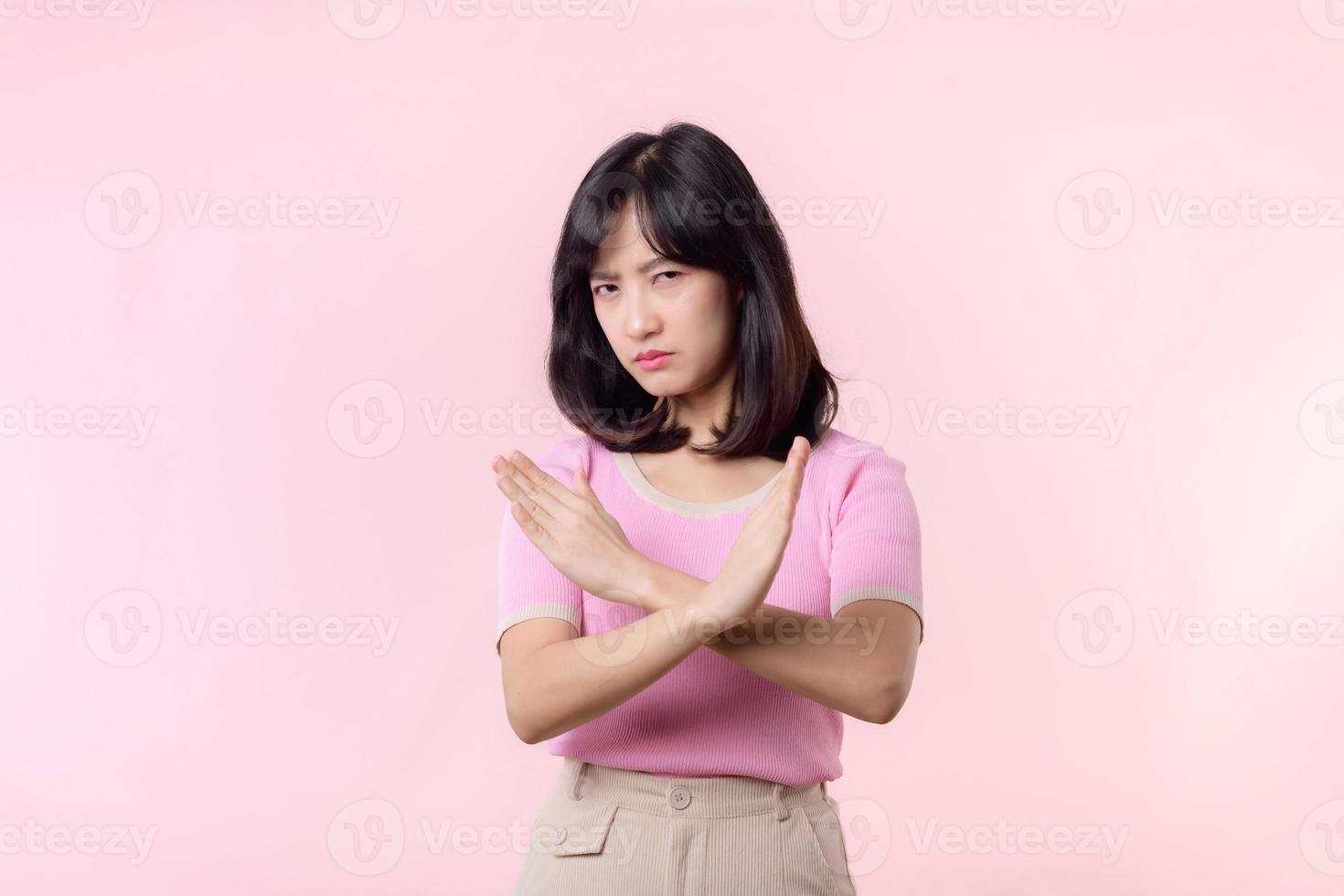 portrait serious young asian woman with cross arm gesture showing stop, no, wrong, denial, rejection sign isolated on pink pastel studio background. deny and negative expression symbol concept. photo