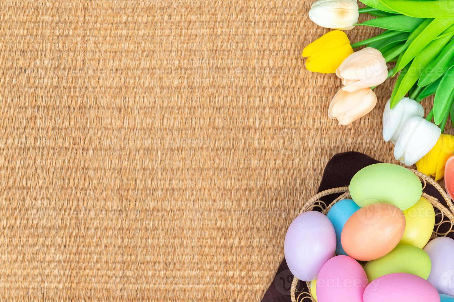 Happy Easter holiday greeting card concept. Colorful Easter Eggs and spring flowers on Papyrus rattan weave texture background. Flat lay, top view, copy space. photo