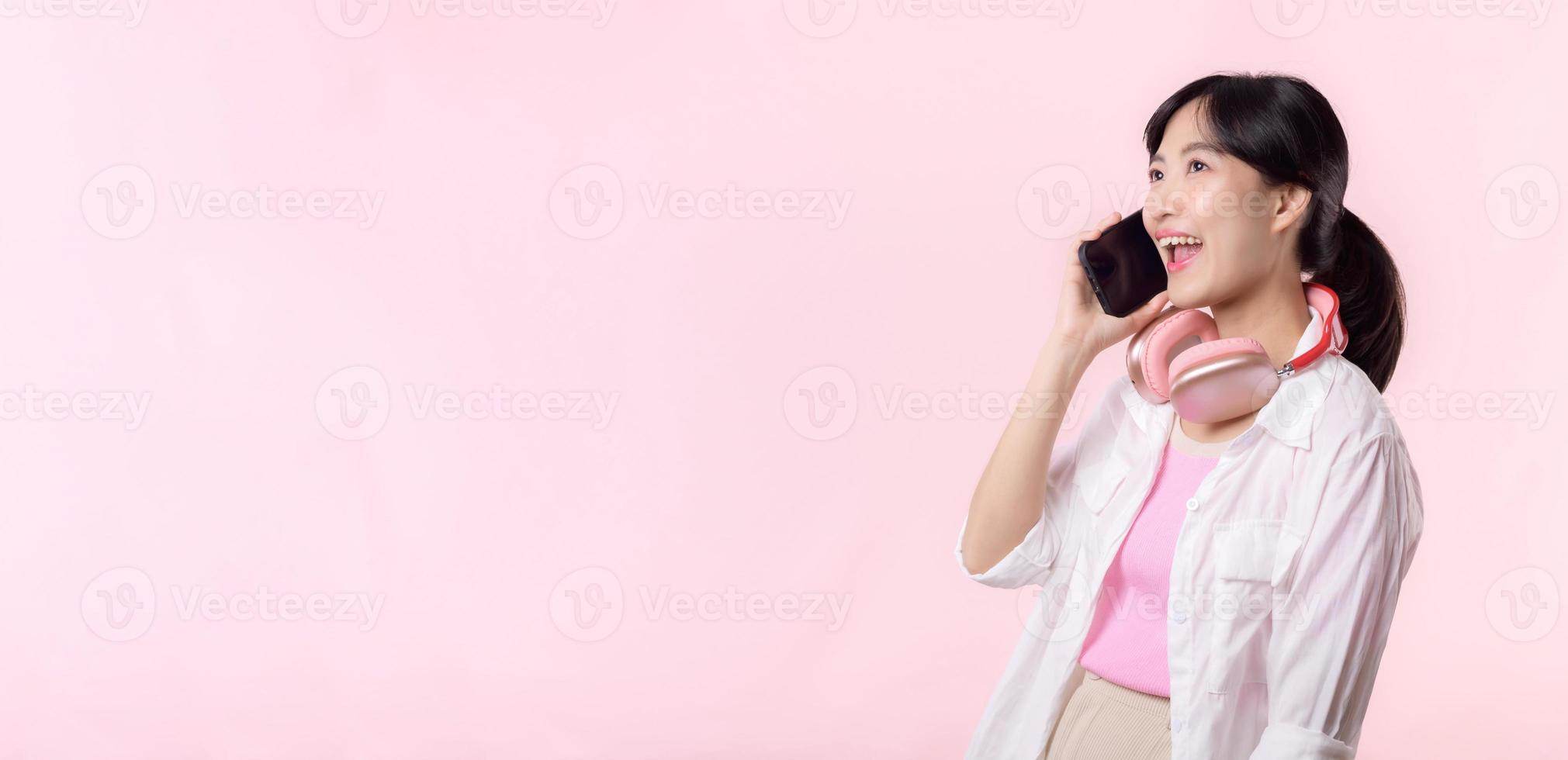 retrato joven atractivo asiático mujer contento sonrisa utilizando teléfono inteligente con auricular, auricular aislado en rosado estudio antecedentes. bonito hembra persona utilizando móvil teléfono. música en línea estilo de vida concepto. foto