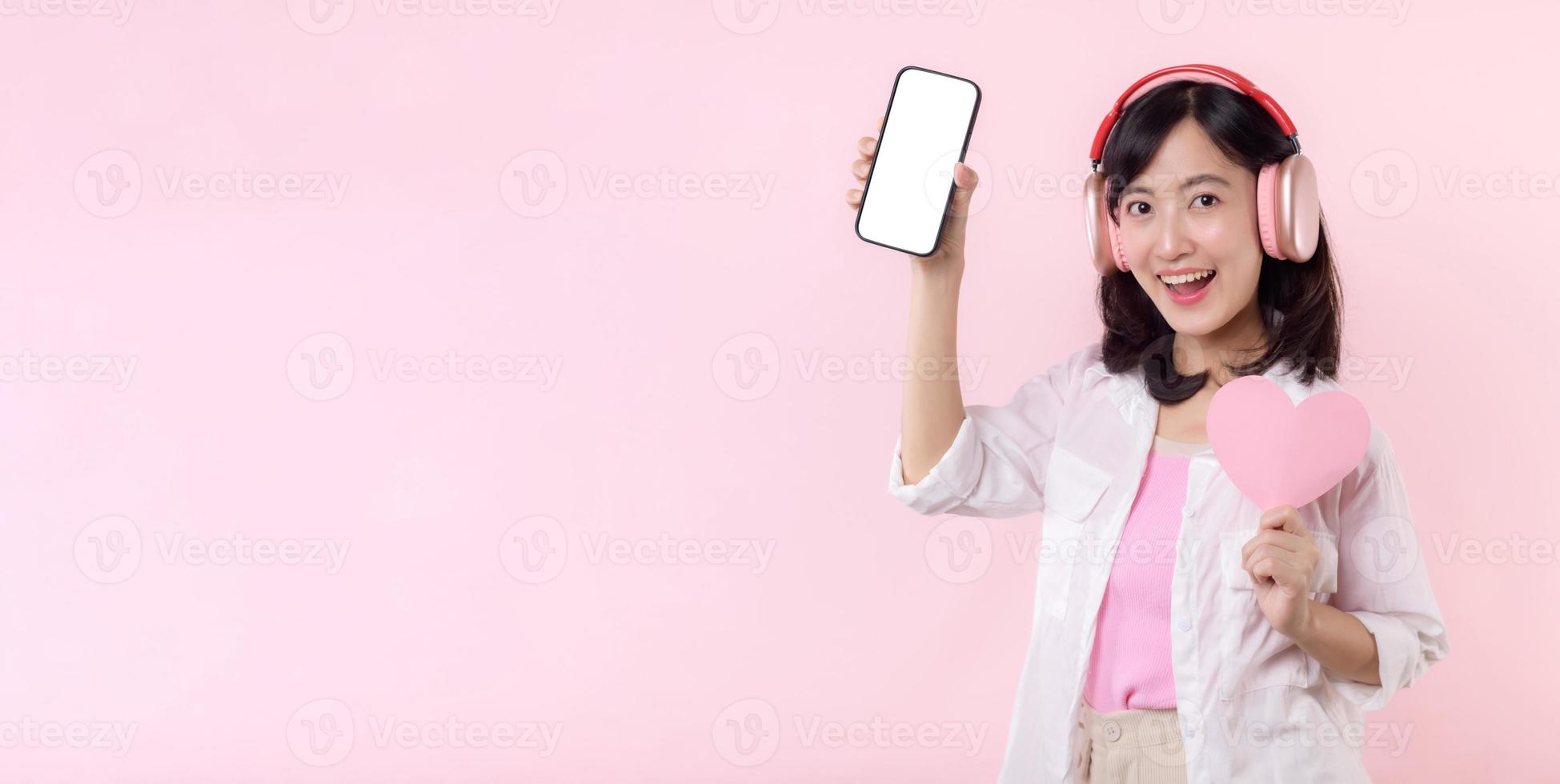 contento alegre sonriente asiático mujer con mano participación corazón y auriculares demostración blanco pantalla móvil teléfono o nuevo teléfono inteligente música solicitud anuncio Bosquejo aislado en rosado estudio antecedentes. foto