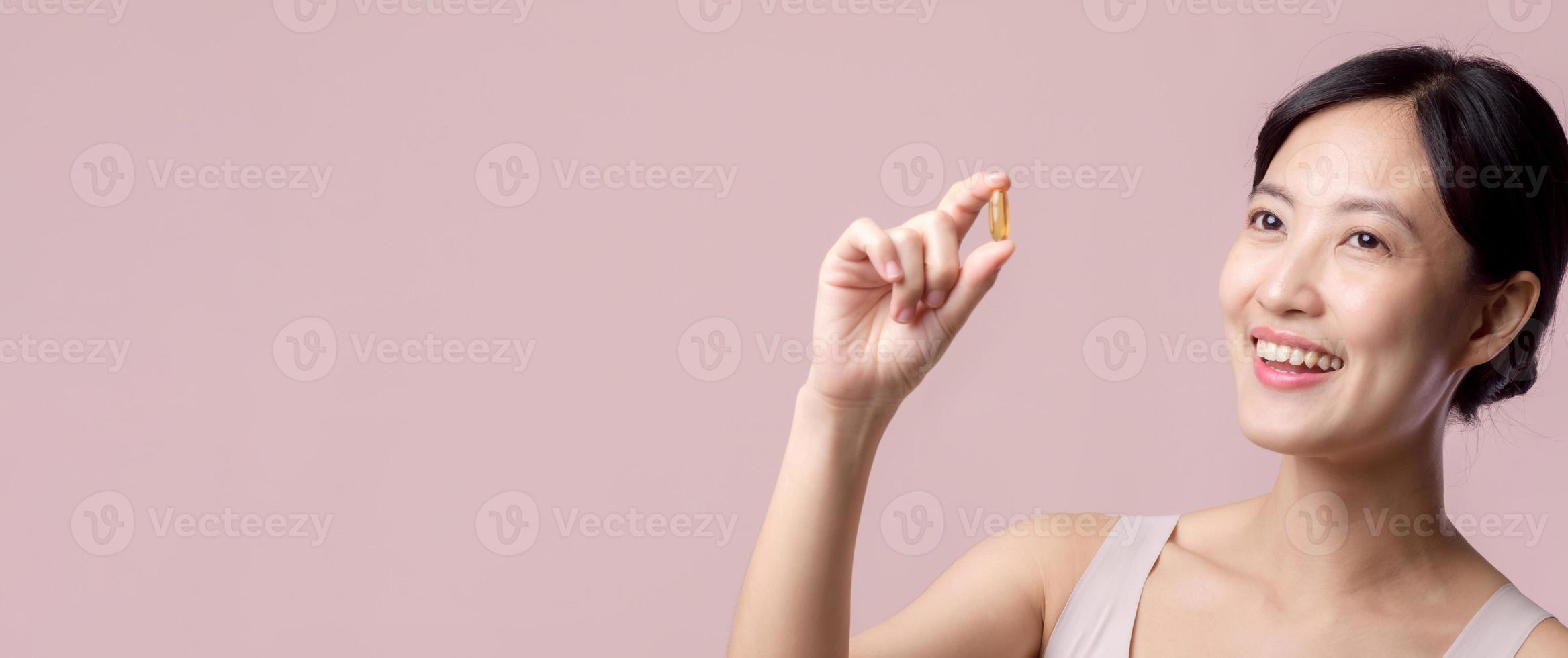 Portrait young asian woman happy smile face with vitamin nourishment pill. Pretty cute girl female person holding health capsule supplement skin care isolated on pink background. Medication concept. photo