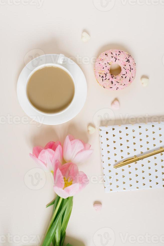 Spring blog concept. Cup of cofee, donut and bouquet of pink tulips on beige background. Flat lay, top view feminine desk. photo