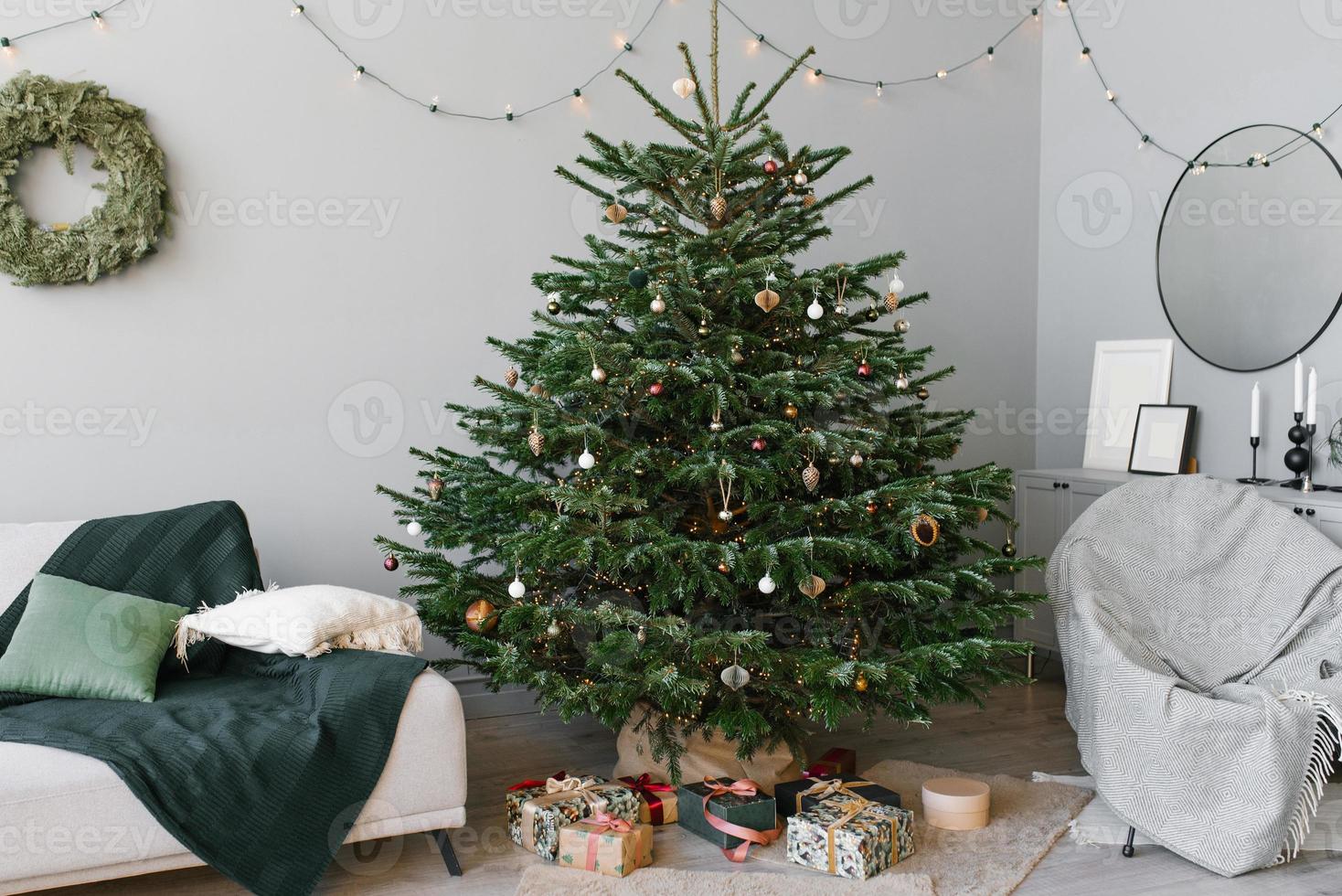 A beautiful Christmas tree fir with toys and a sofa with pillows in the interior of the living room in the Scandinavian style with gray walls photo