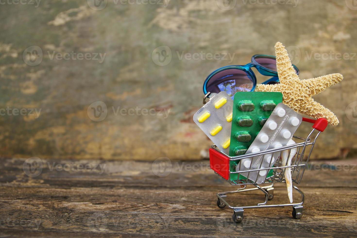 Shopping cart with pills, sunglasses, seashells on old wooden background. Concept of medication required in journey. photo