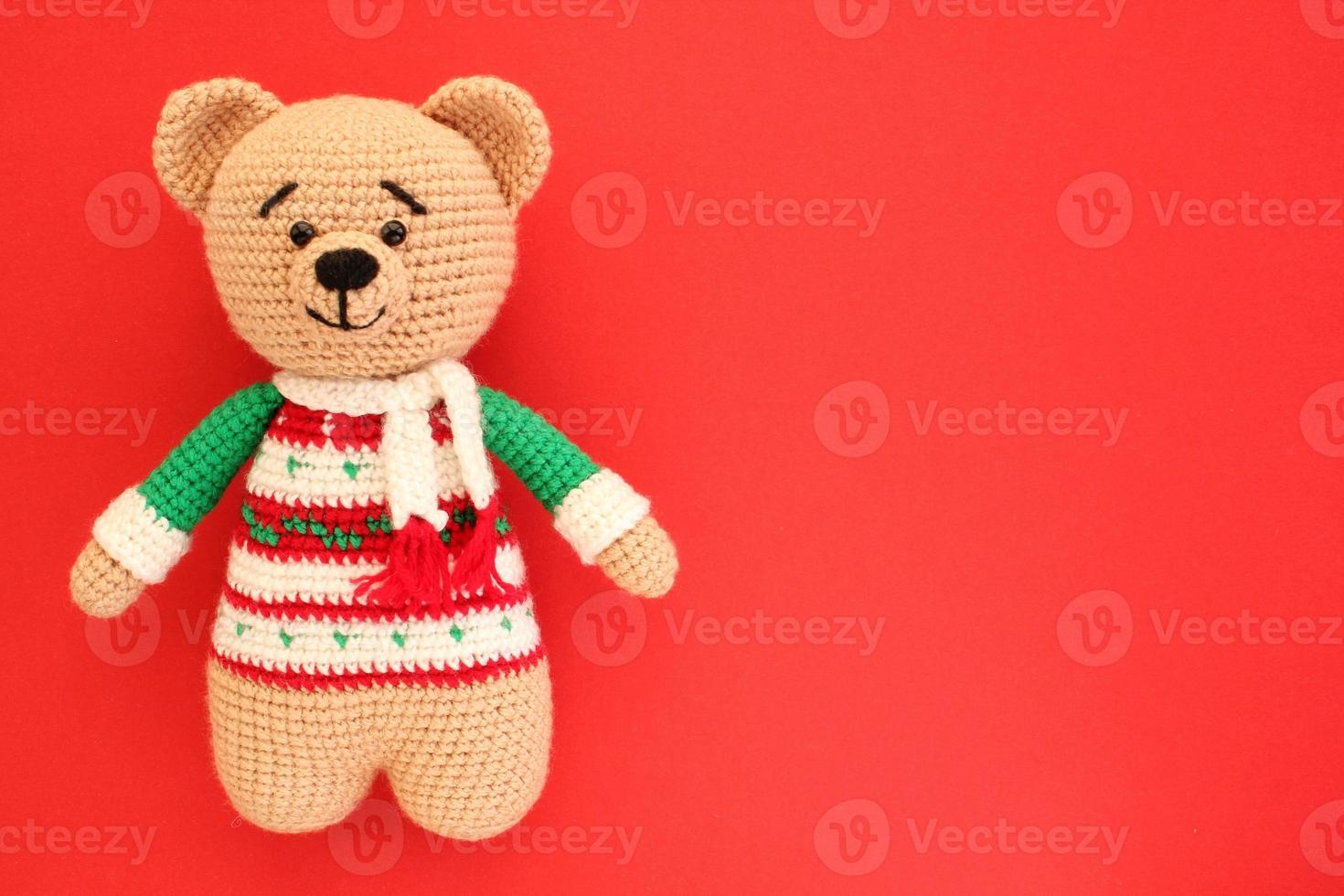 Crochet amigurumi handmade stuffed soft teddy bear toy in colored sweater on red background. Handwork, hobby. Craft diy newborn pregnancy concept. Knitted doll for little baby. Closeup flat lay out photo