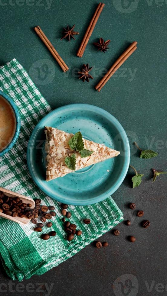 parte superior ver de delicioso Napoleón pastel con crema en un turquesa plato decorado con un puntilla de menta en un verde antecedentes. un taza de caliente café, canela palo, badyan, café frijoles en un verde fondo foto