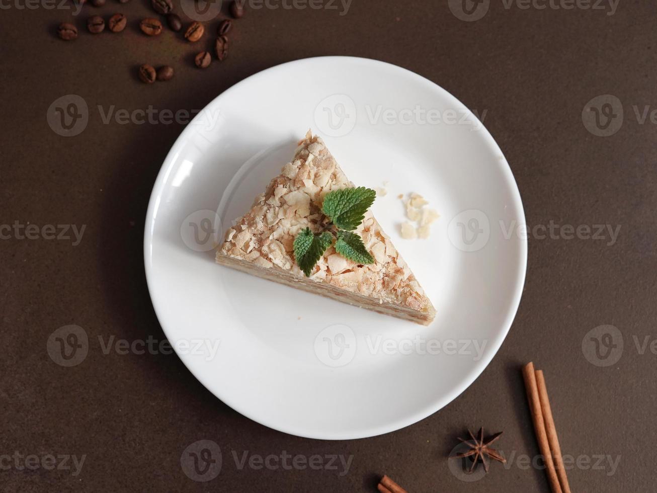 Top view of Delicious Napoleon Cake with cream on a white plate decorated with a sprig of mint on a Brown Background. A cinnamon stick, badyan, coffee beans on a brown background. Copy space photo