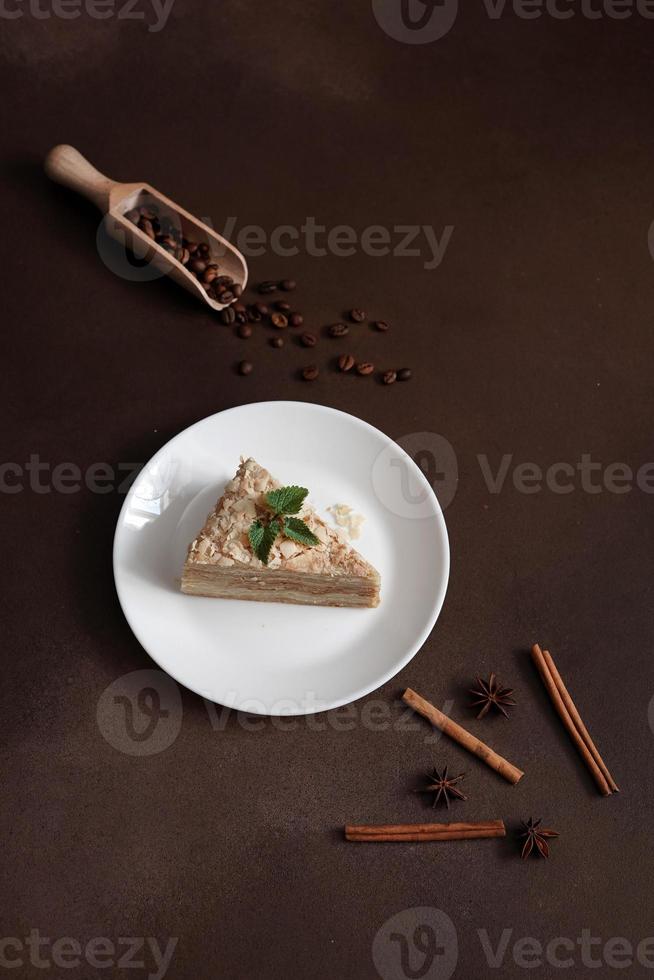 Delicious Napoleon Cake with cream on a white plate decorated with a sprig of mint on a Brown Background. A cinnamon stick, badyan, coffee beans on a brown background. Copy space photo