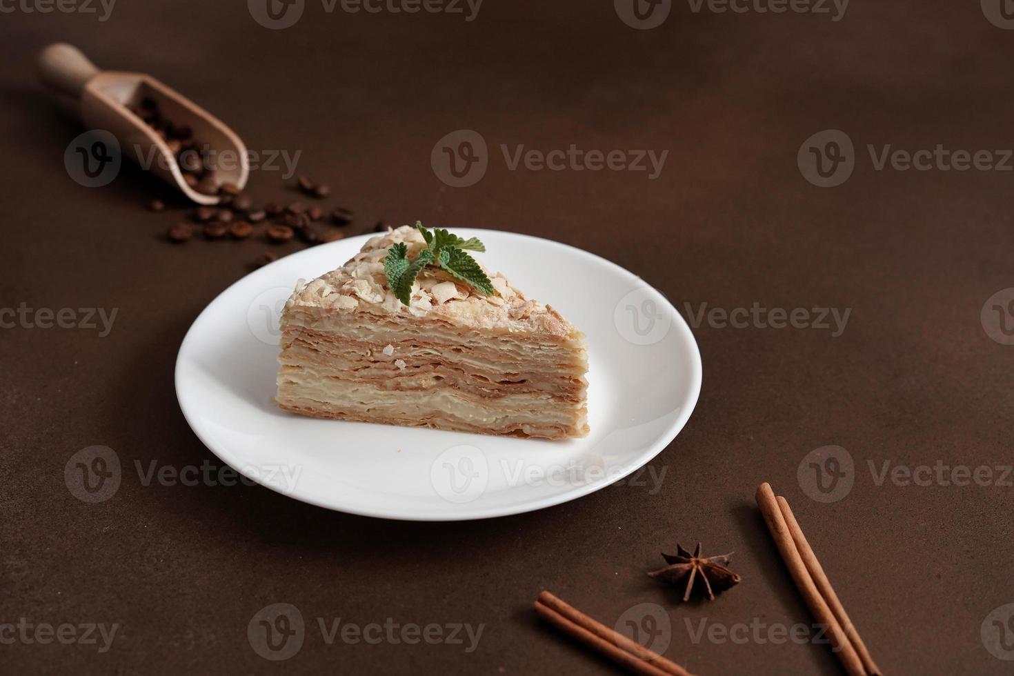 Delicious Napoleon Cake with cream on a white plate decorated with a sprig of mint on a Brown Background. A cinnamon stick, badyan, coffee beans on a brown background. Copy space photo