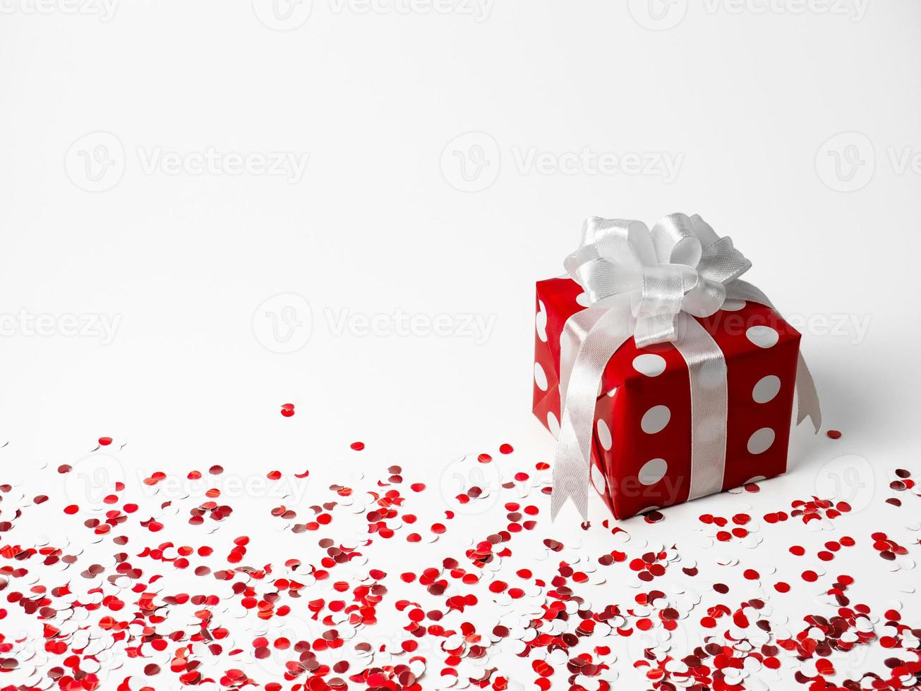 gift for New Year, Christmas, Birthday, loved ones. Red polka dot box with white bow on white background with confetti. photo