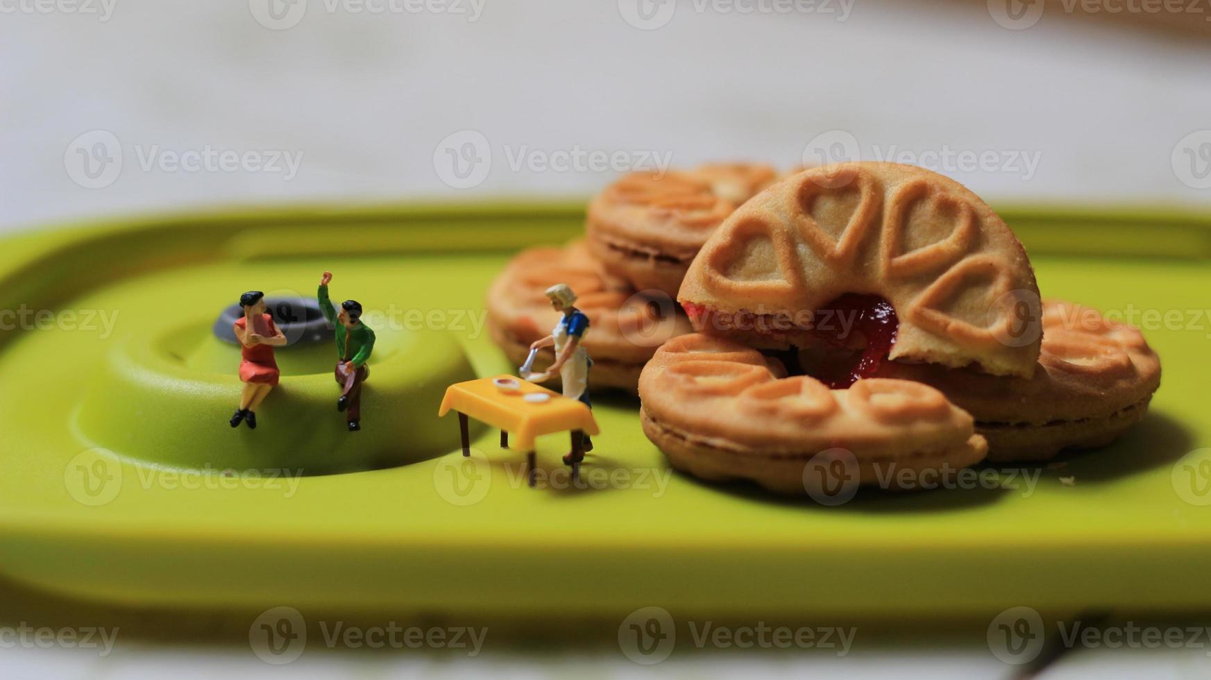 Miniature figures of a couple talking on a green dining table near a chef cooking on a background of a cake filled with strawberry jam. discussion concept. photo