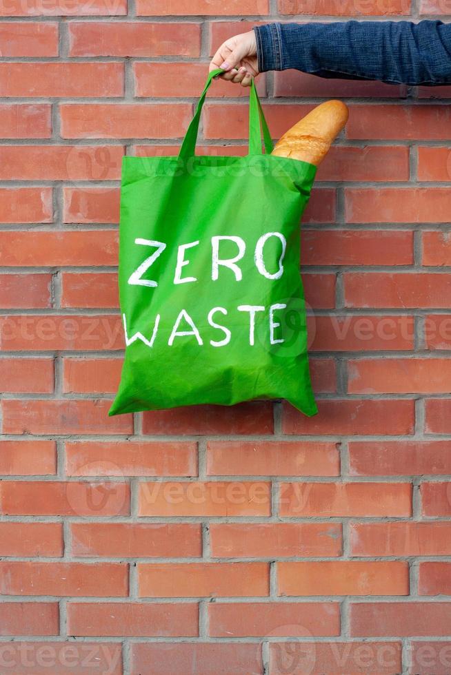 Green eco bag with a white text zero waste in the hand on a background of a brick wall with copy space. photo