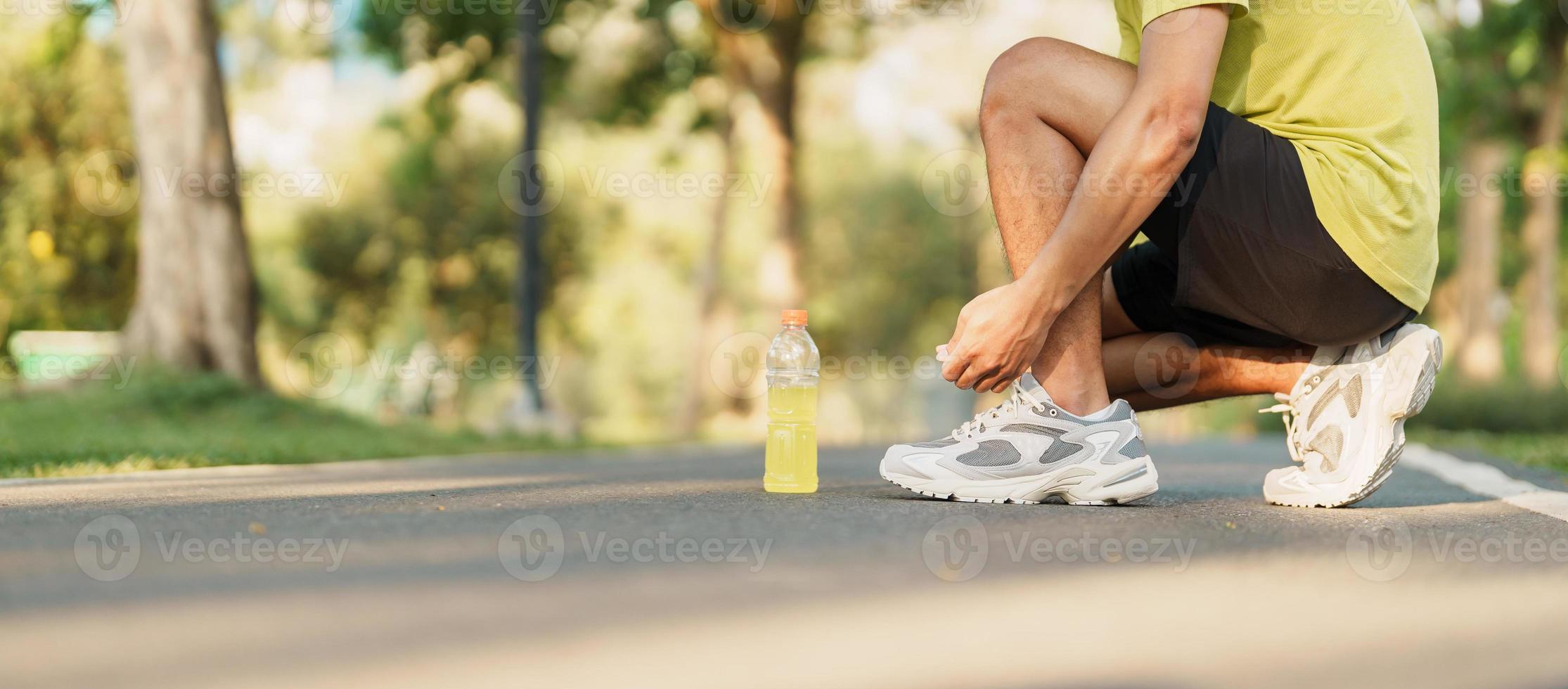 Young athlete man tying running shoes with Energy Drink water, male runner ready for jogging outside, asian Fitness walking and exercise in the park morning. wellness, wellbeing and sport concepts photo