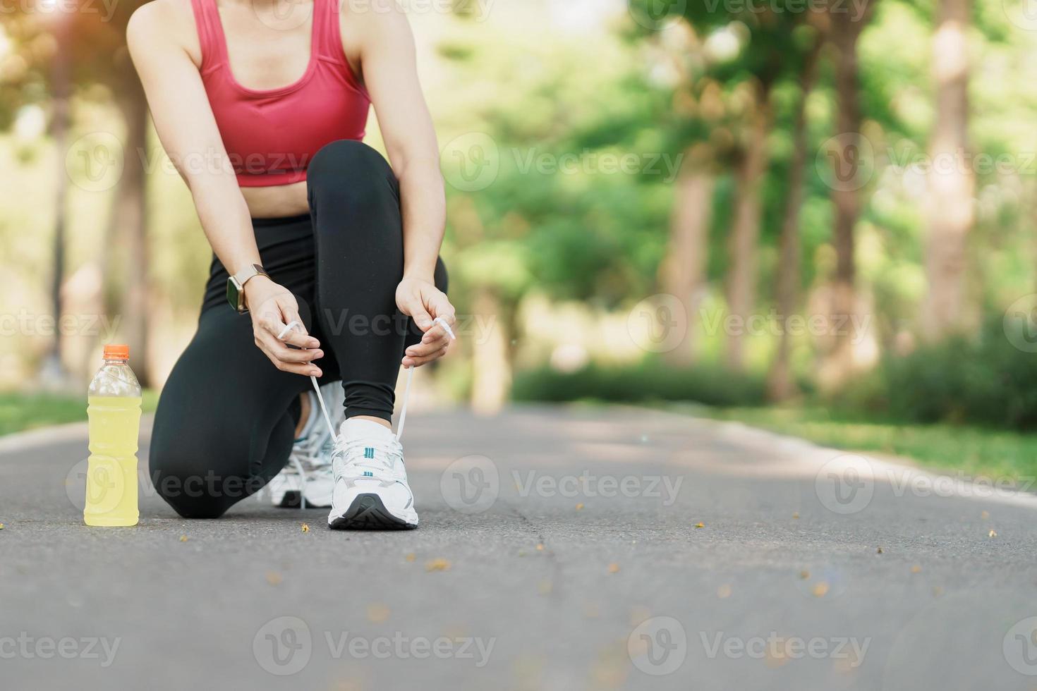 Young athlete woman tying running shoes with Energy Drink water, female runner ready for jogging outside, asian Fitness walking and exercise in the park morning. wellness, wellbeing and sport concepts photo