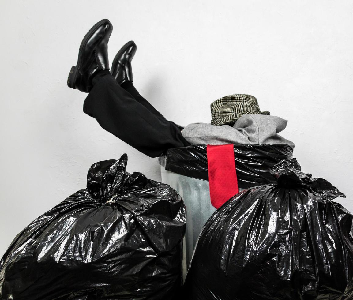 Profile of Businessman in Suit Stuck Inside Metal Trash Can Next to Garbage Bag Pile. Concept of Man Beaten Up and Thrown Away by Capitalism and Greed. photo