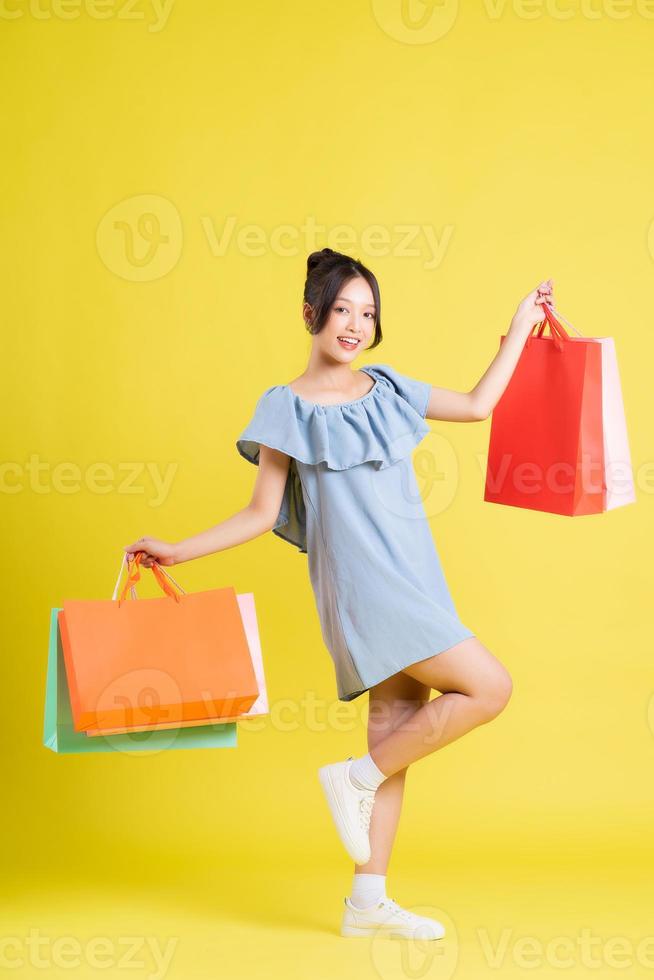 imagen de un hermosa asiático niña en un vestir participación un compras bolso en su mano posando en un amarillo antecedentes foto