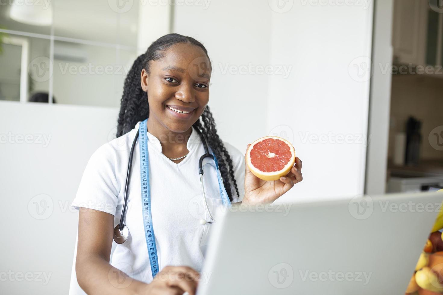 Portrait of young smiling female nutritionist in the consultation room. Nutritionist desk with healthy fruit, juice and measuring tape. Dietitian working on diet plan. photo