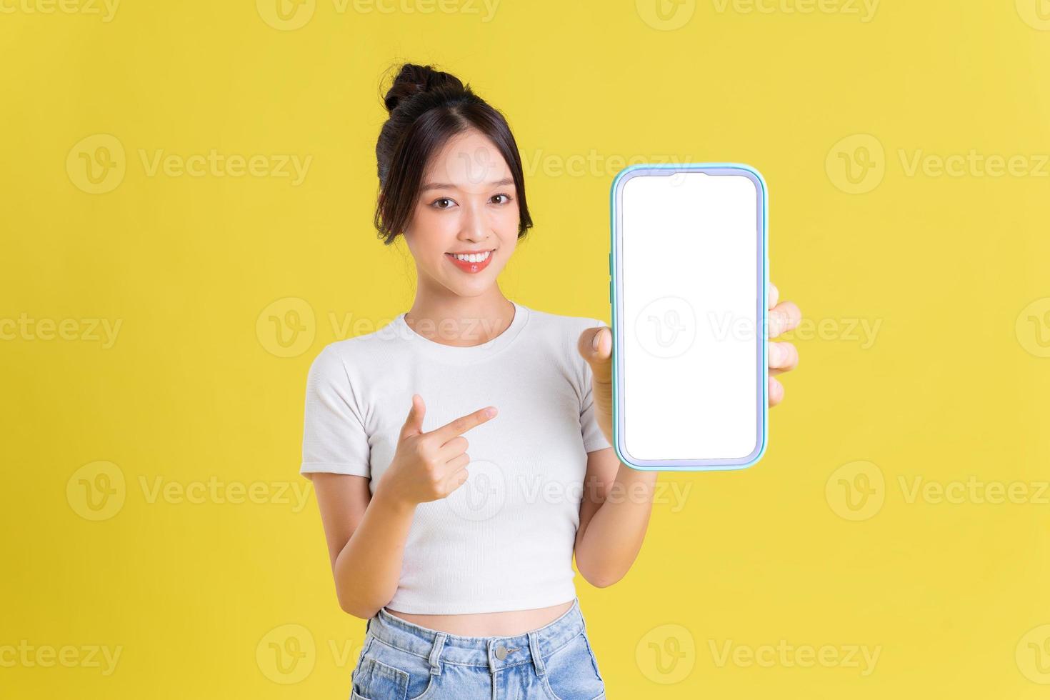 Young Asian woman holding phone with cheerful face on yellow background photo