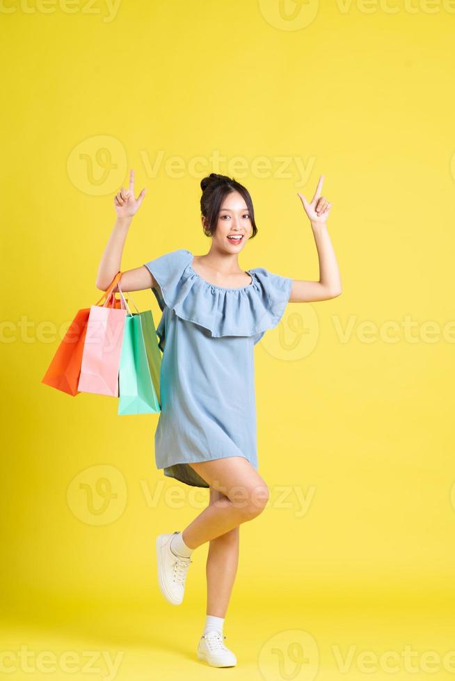 imagen de un hermosa asiático niña en un vestir participación un compras bolso en su mano posando en un amarillo antecedentes foto