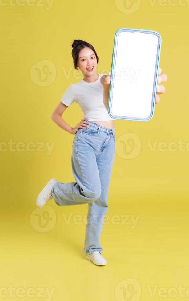 full body image of young Asian girl holding phone with cheerful face on yellow background photo