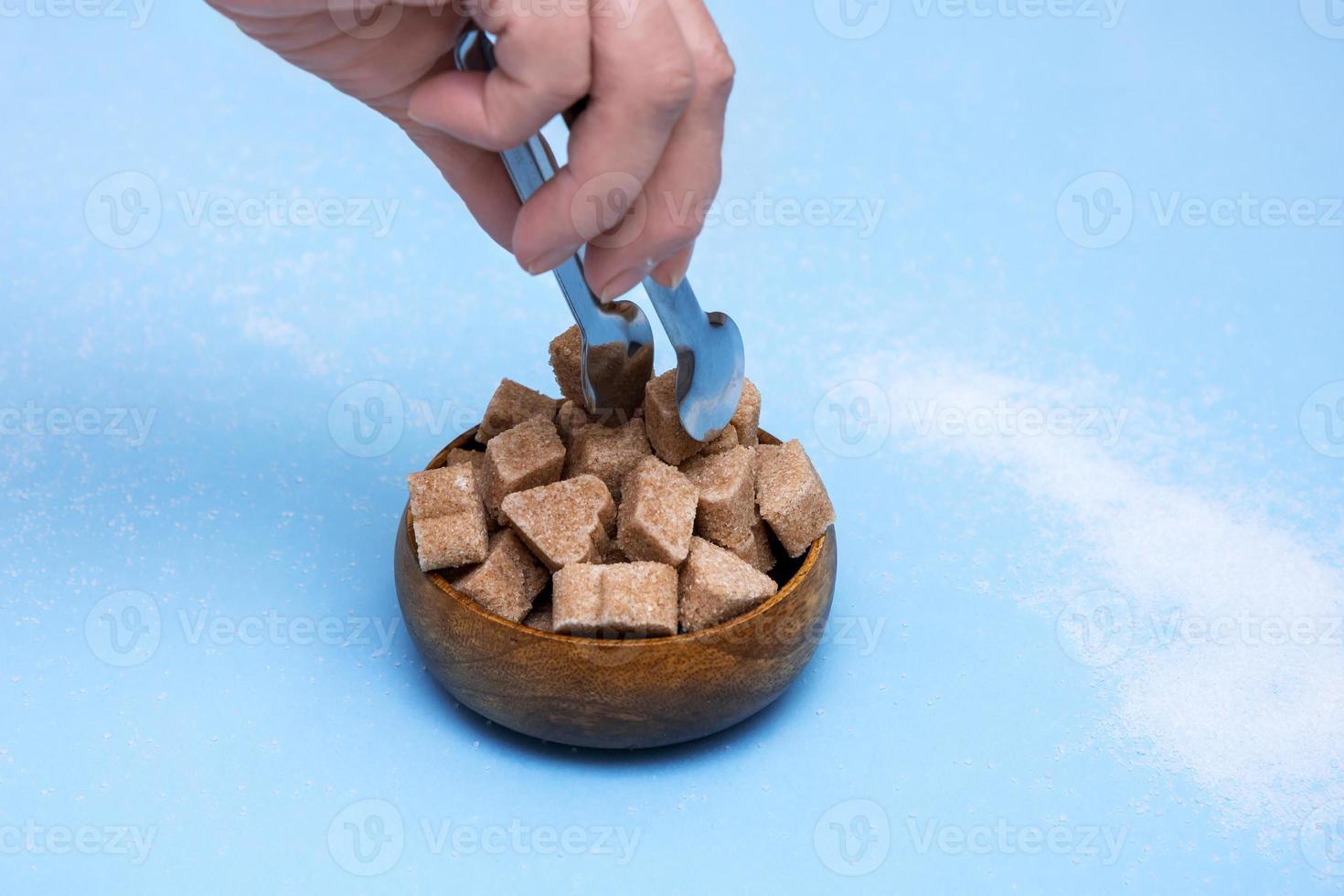 hand takes with tongs lump sugar with cinnamon from a bowl on a blue background photo