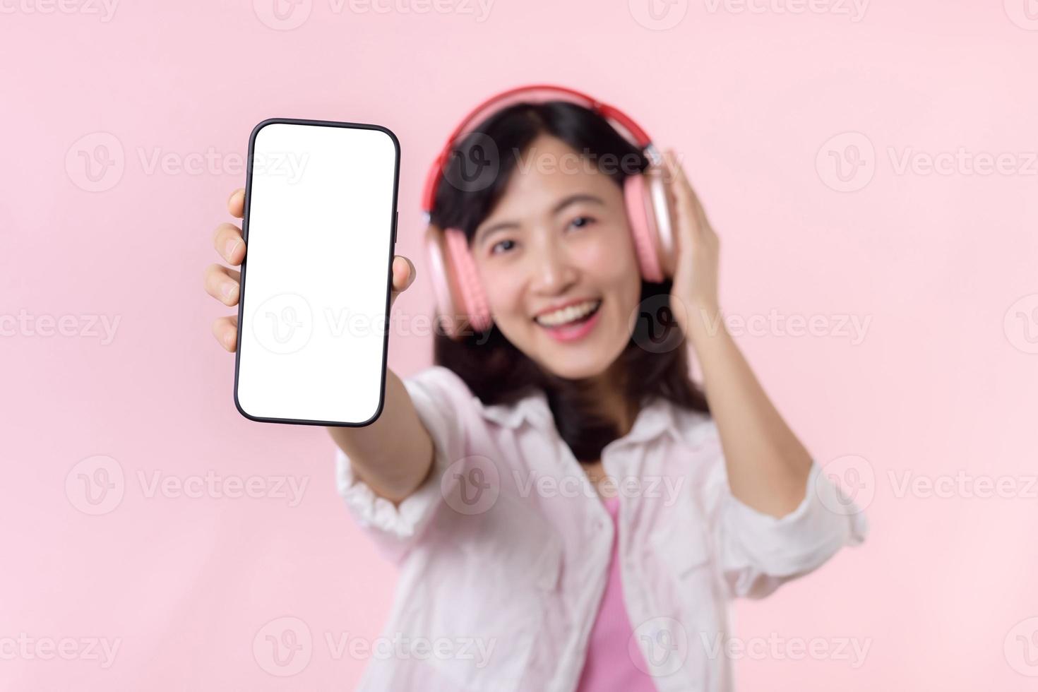 Happy cheerful smiling asian woman with wireless earphones showing blank screen mobile phone or new smartphone music application advertisement mockup isolated on pink studio background. photo