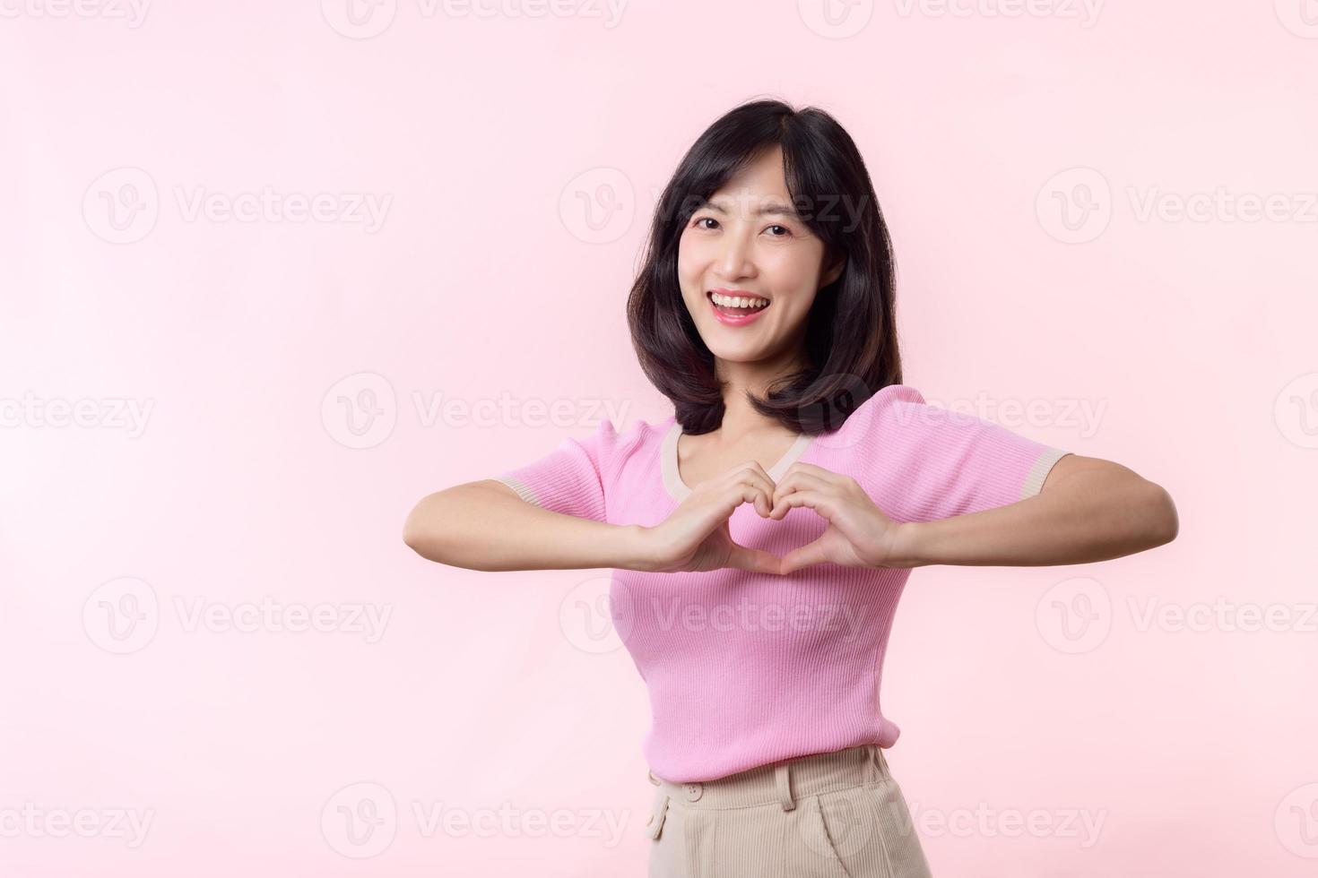 portrait young asian woman feels happy, love and romantic shapes heart hand gesture expresses tender feelings wears pink casual shirt against pastel pink background. People affection and care concept photo