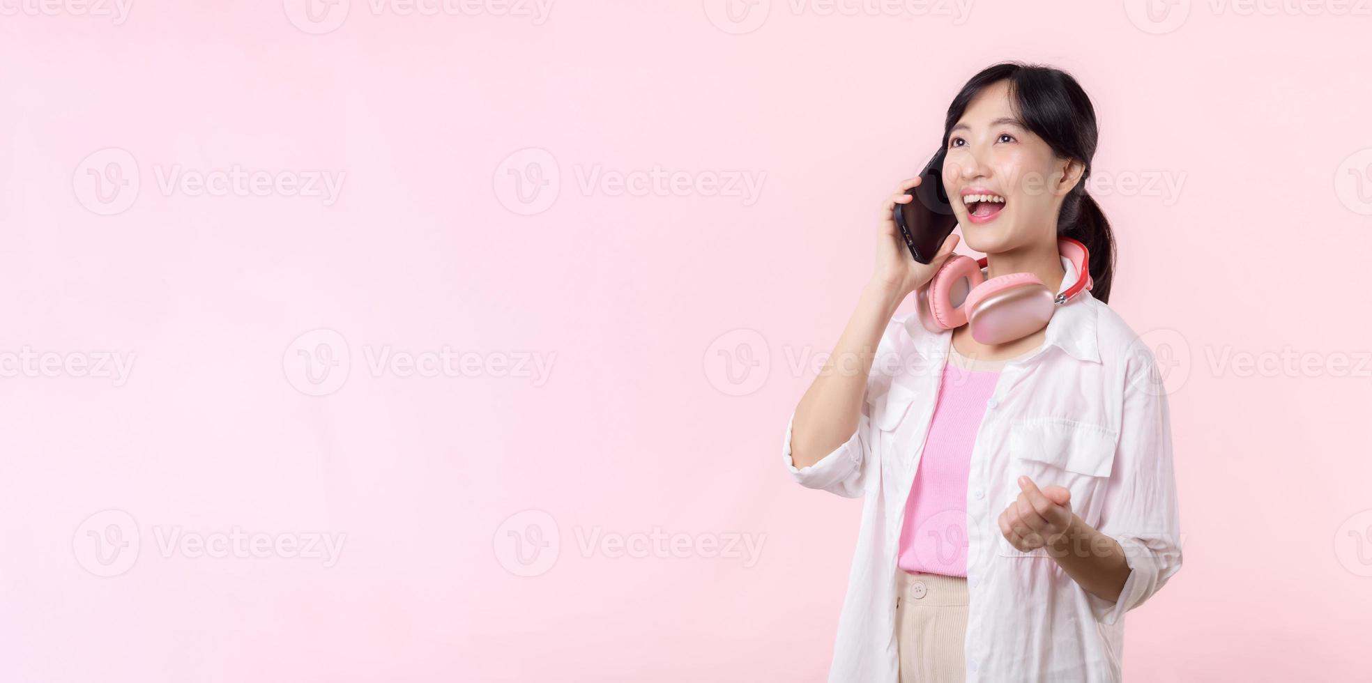 retrato joven atractivo asiático mujer contento sonrisa utilizando teléfono inteligente con auricular, auricular aislado en rosado estudio antecedentes. bonito hembra persona utilizando móvil teléfono. música en línea estilo de vida concepto. foto