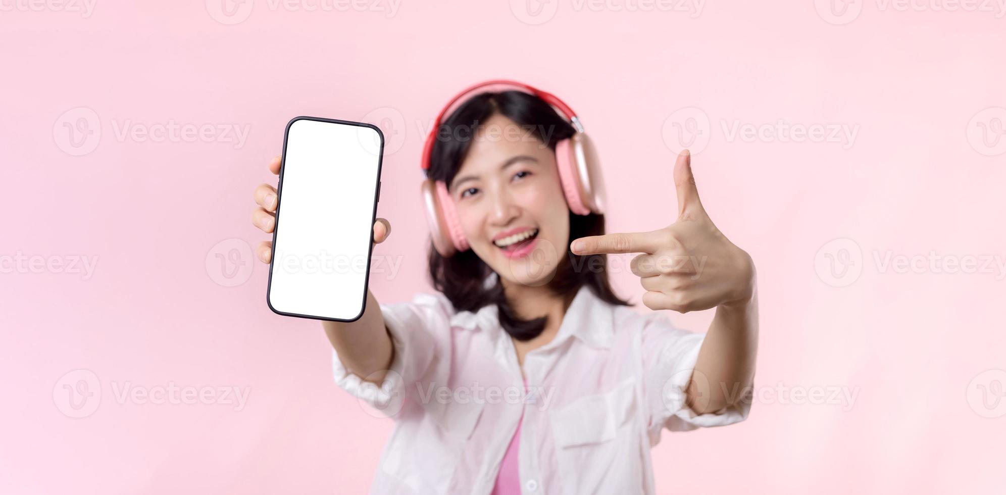 contento alegre sonriente asiático mujer con inalámbrico auriculares demostración blanco pantalla móvil teléfono o nuevo teléfono inteligente música solicitud anuncio Bosquejo aislado en rosado estudio antecedentes. foto