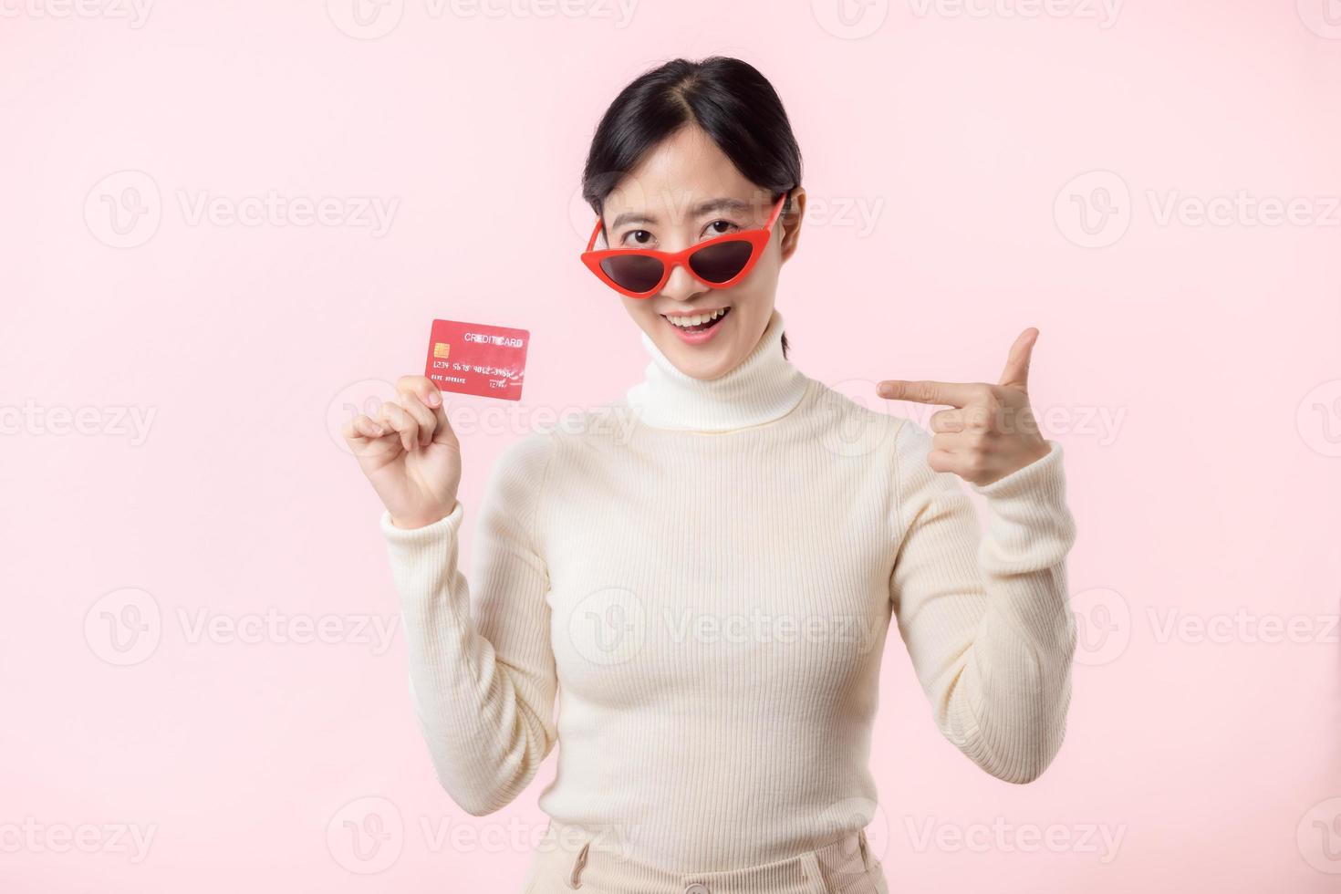 fascinante divertido alegre joven mujer de asiático etnia 20s años antiguo con vestir Gafas de sol usa blanco camisa sostener en mano crédito banco tarjeta aislado en llanura pastel ligero rosado antecedentes estudio retrato. foto