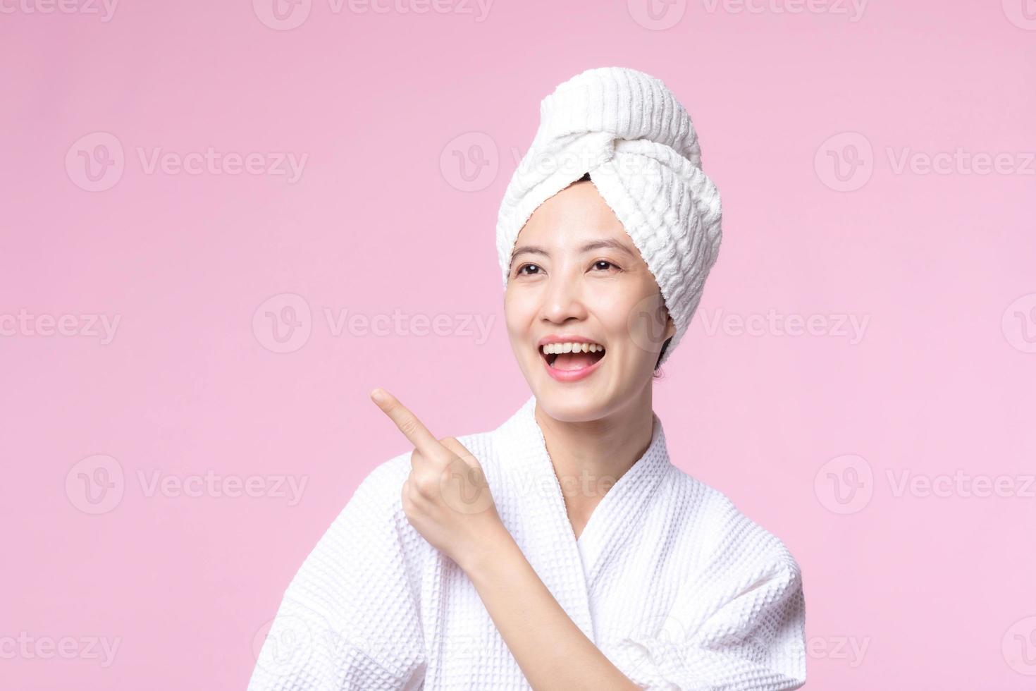 Beautiful young asian woman happy smile face with hand or finger gesture signs in bathrobe and shower towel showing empty space isolated on pink background. photo