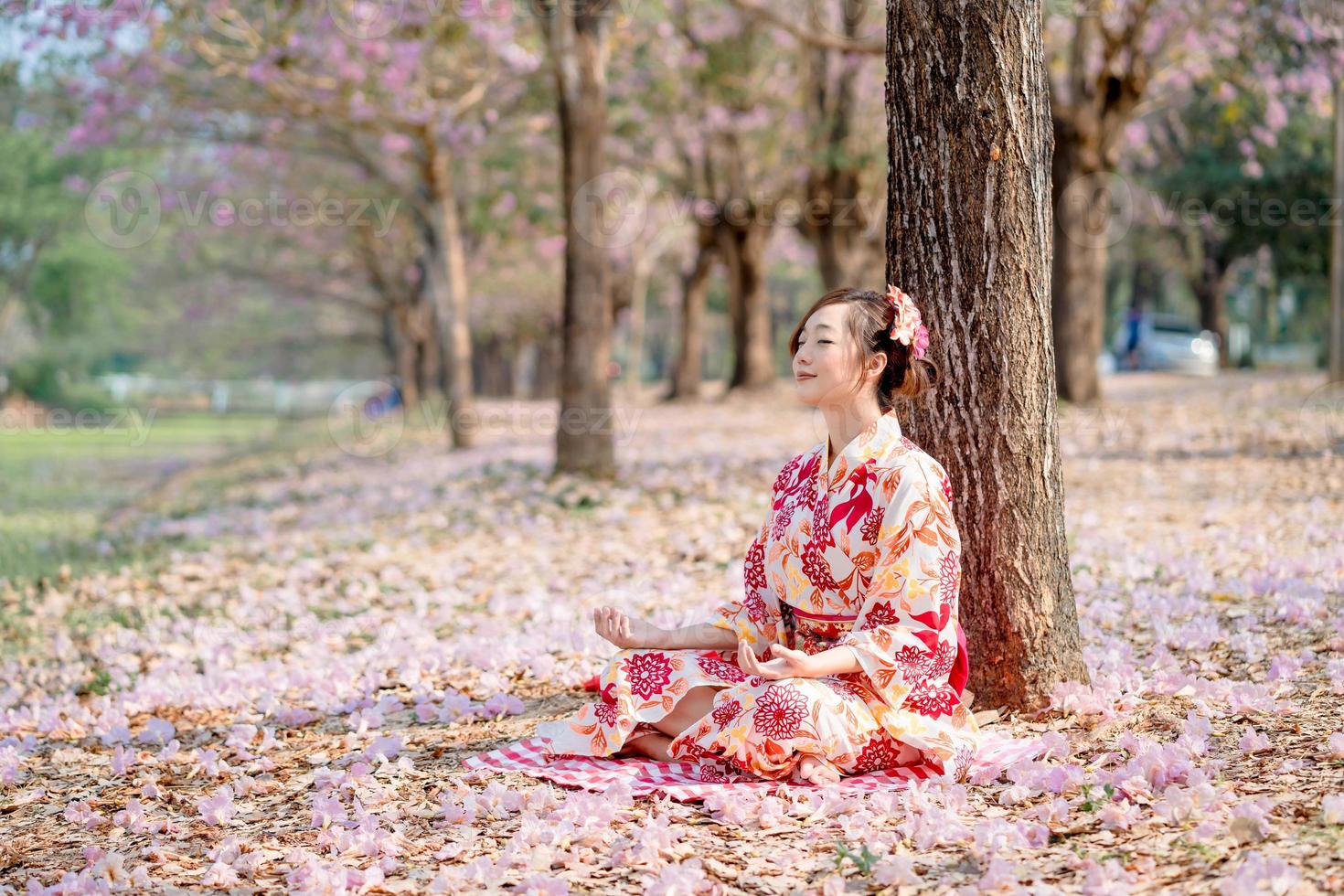 Young lady in traditional kimono dress practices meditation at cherry blossom tree. Emotion smile photo