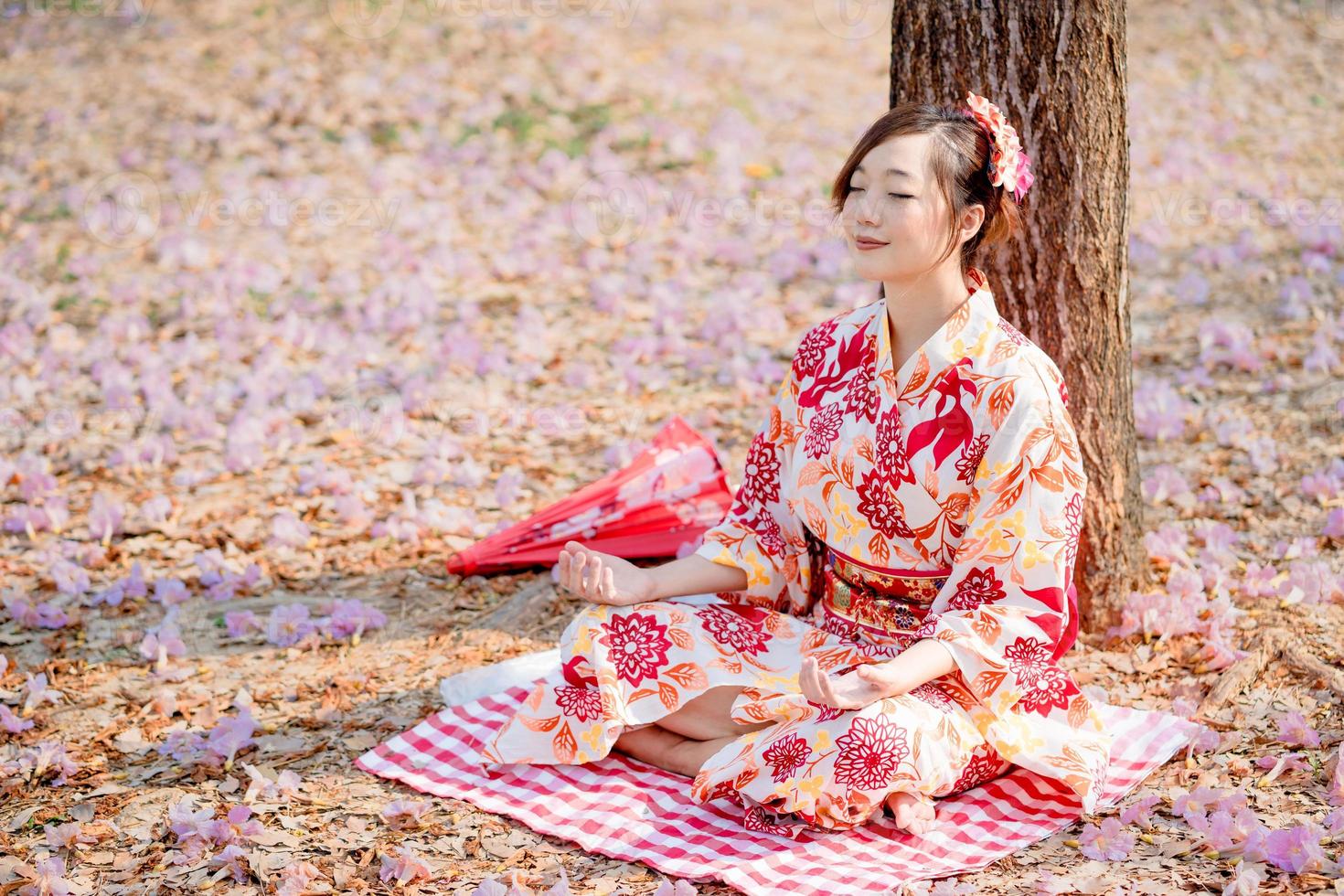 Asian woman in traditional kimono dress practices meditation at cherry blossom tree. photo