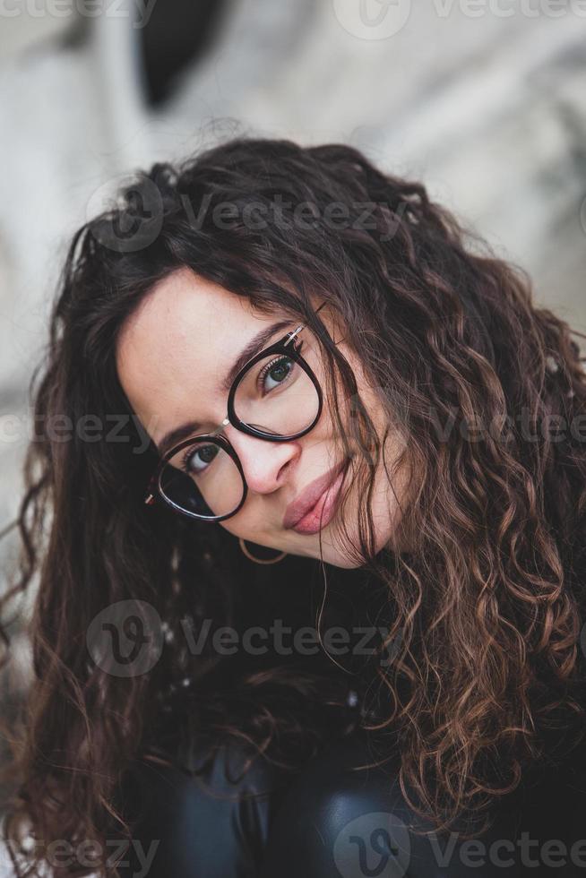 Beautiful young woman with brunette curly hair, portrait in eye glasses enjoying the sun in the city. photo
