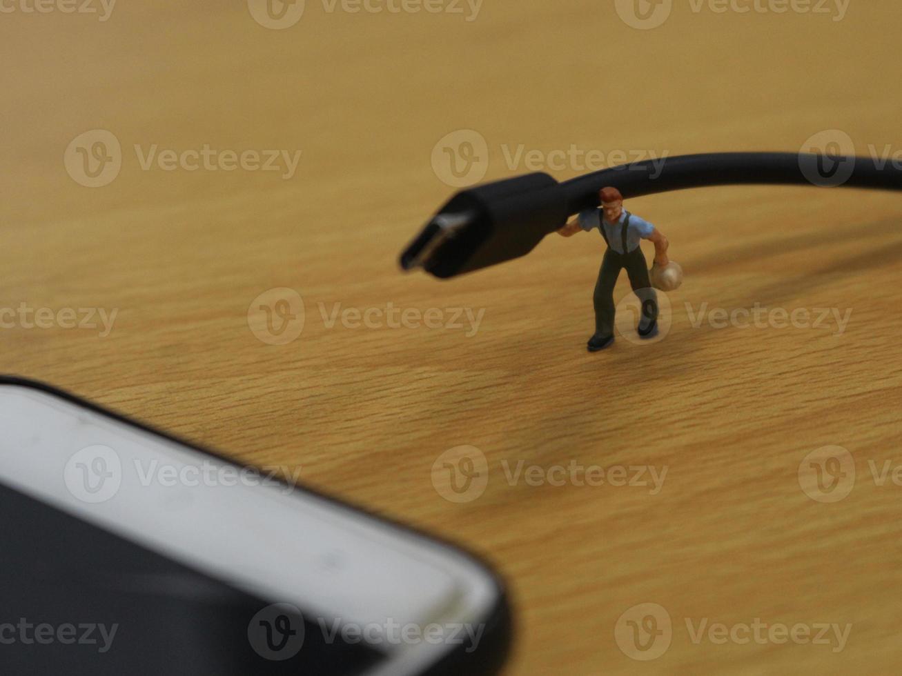 un miniatura figura de un trabajador que lleva un Teléfono móvil cargando cable. foto