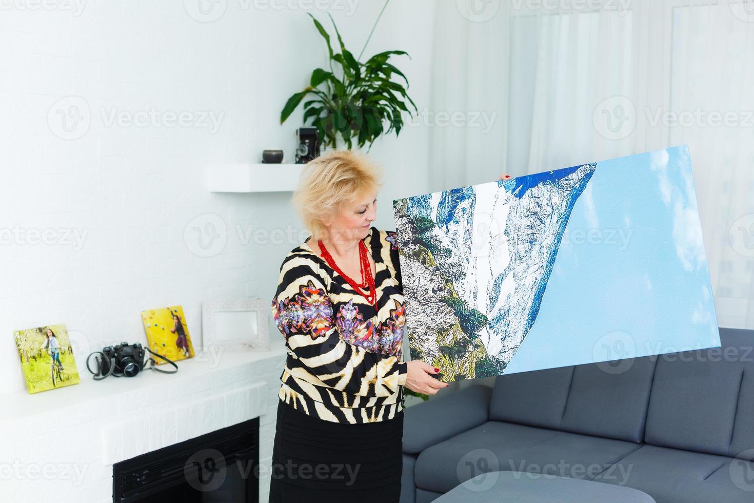 A happy elderly blonde woman is holding a large wall photo canvas at home