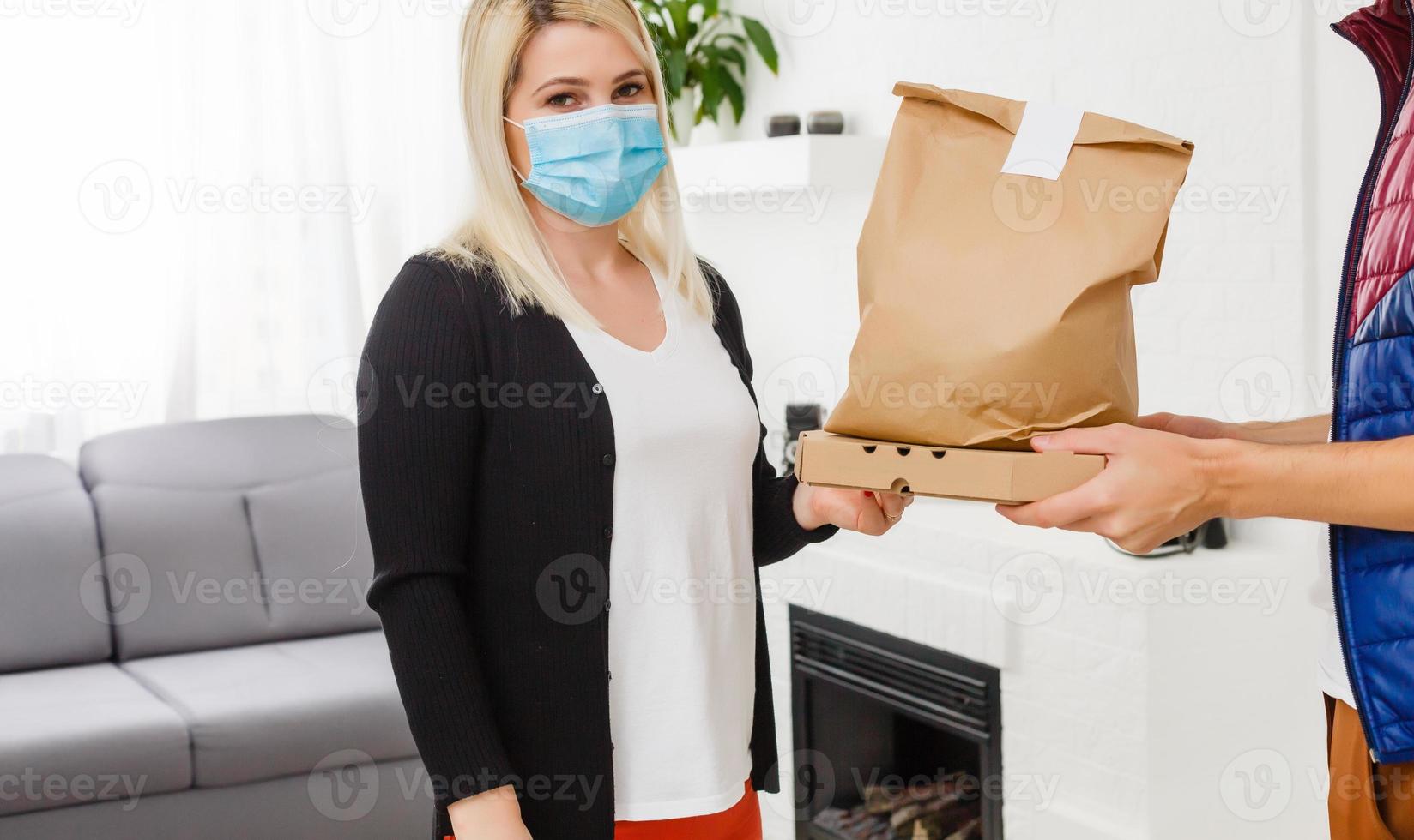 Young woman wearing medical mask receiving parcel from delivery man indoors. Prevention of virus spread photo