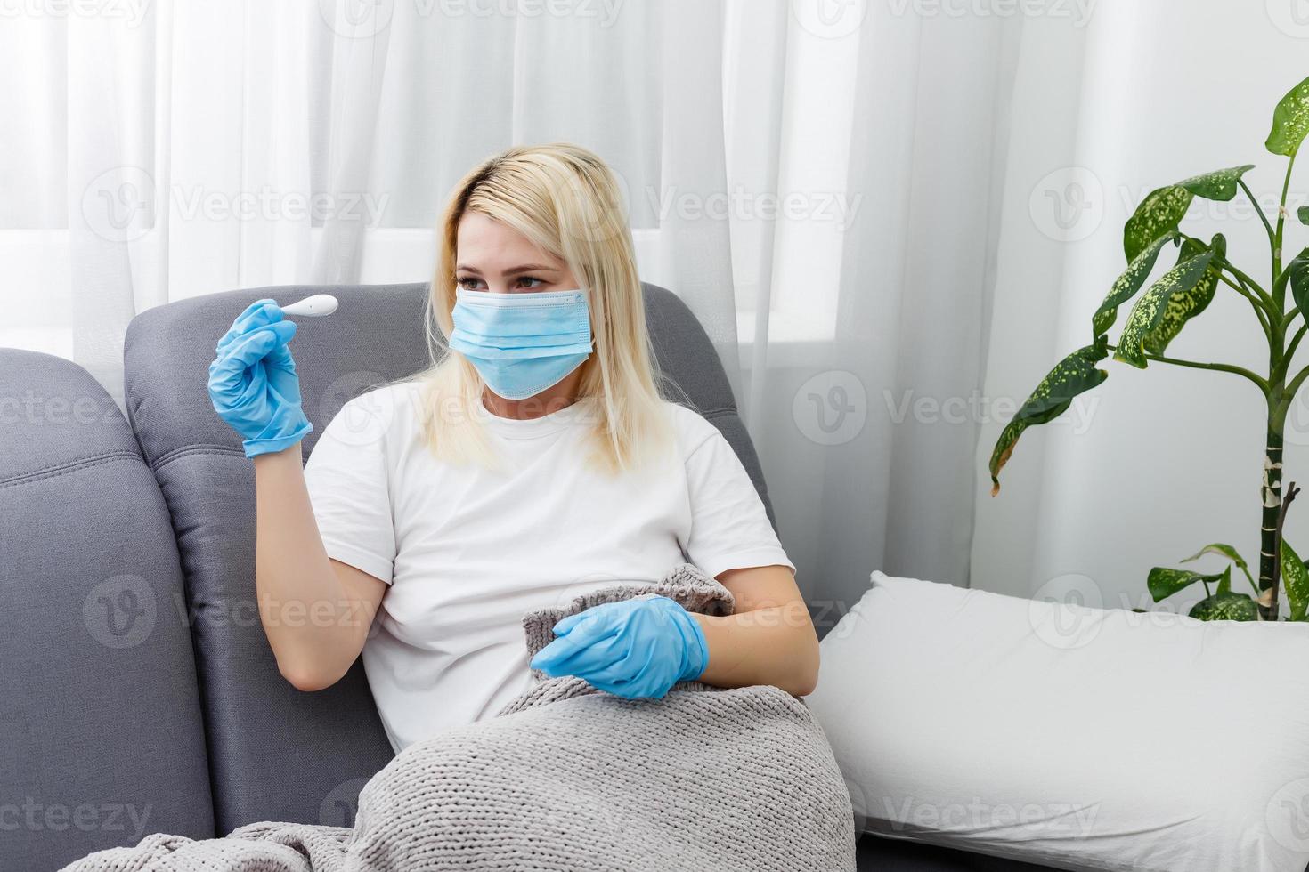 Fever and coronavirus symptoms, woman in medical mask measures body temperature. Sick girl looks at digital thermometer in her hands, concept of cold and flu photo