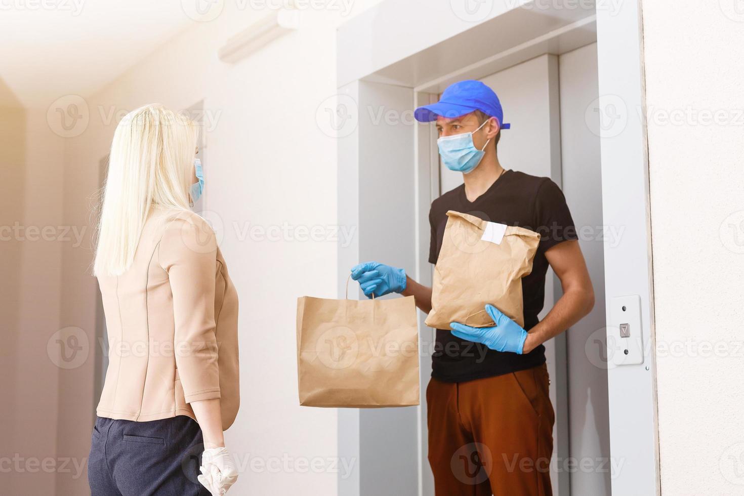 Young woman wearing medical mask receiving orderd food from the restaurant from delivery man indoors. Prevention of virus spread photo