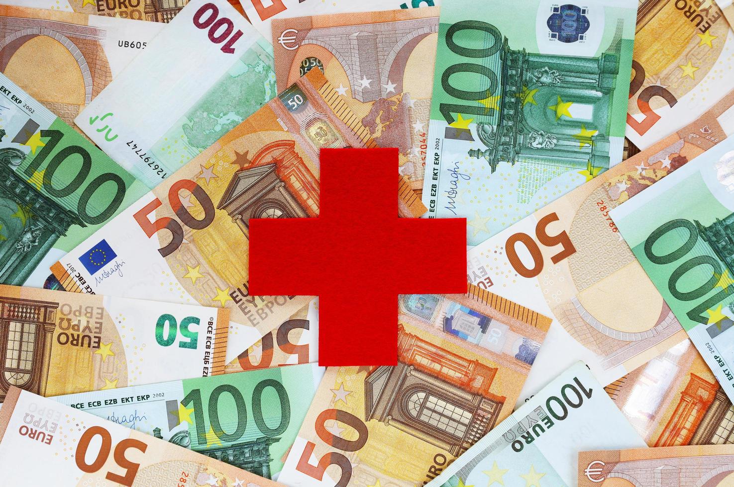 KRAKOW, POLAND, MARCH, 2021 - Medical cross symbol cut out of red felt centered on background with 50 and 100 euro banknotes. Healthcare system in Europe. Paid medicine. Costs, treatment prices. photo